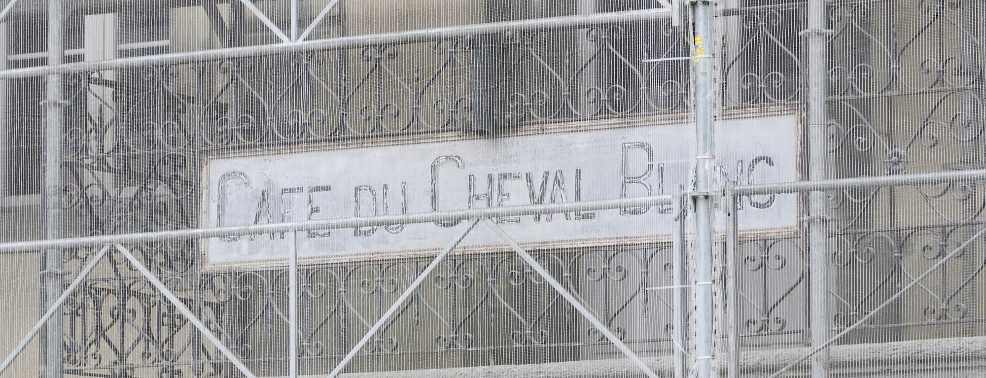 L'enseigne gravée « Café du Cheval Blanc » et le balcon en fer forgé, aperçus derrière l'échafaudage pendant la rénovation